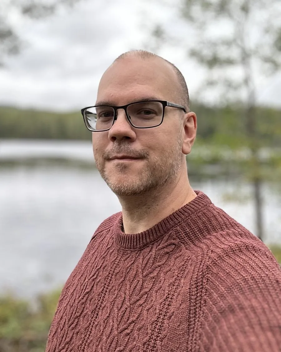 Björn Sennbrink by a lake in the woods, in glasses and a cable-knit sweater, looking into the camera.
